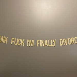 May include: Gold glitter letters spelling out the phrase "Thank Fuck I'm Finally Divorced" on a gray background.