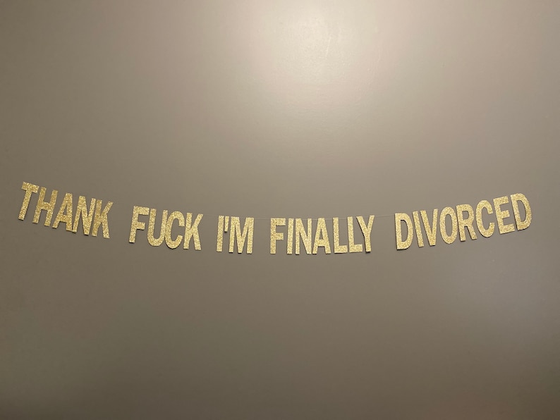 May include: Gold glitter letters spelling out the phrase "Thank Fuck I'm Finally Divorced" on a gray background.