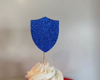 May include: A white cupcake with white frosting topped with a blue glitter shield cupcake topper.