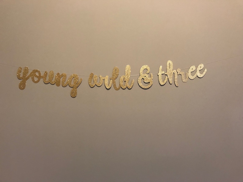 Puede incluir: Pancarta de purpurina dorada con el texto "young wild & three".