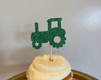 May include: Green glitter tractor cupcake topper. The tractor is made of green glitter paper and has a black outline. The topper is on a white cupcake with vanilla frosting.