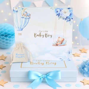 May include: A light blue gift box with a matching bow, featuring a decorative lid with a baby boy theme. The lid displays a hot air balloon, a teddy bear on a swing, and the text "Our Little Baby Boy". A small bag with "Precious Keepsakes" is also present.