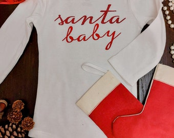 May include: White long-sleeved baby onesie with red glitter lettering that says "santa baby". A red and white stocking is also in the image.