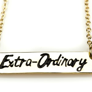 May include: A gold chain necklace with a gold bar pendant that says "Extra-Ordinary" in black cursive lettering.