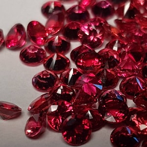 May include: A collection of loose red gemstones, likely rubies, with a faceted cut. The gemstones are scattered on a white surface.