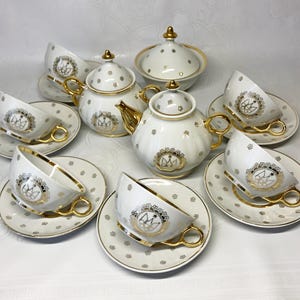 Puede incluir: Un juego de té de porcelana blanca con detalles dorados. El juego incluye una tetera, azucarero y varias tazas y platillos. Cada pieza está decorada con un emblema dorado y pequeños motivos florales. Las tazas tienen asas y bordes dorados.