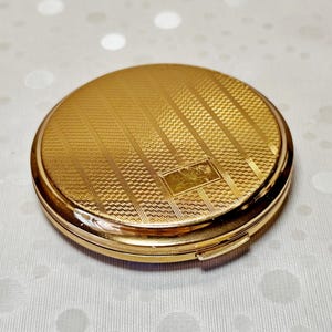 May include: A round gold-tone metal compact mirror featuring a textured surface with alternating diagonal stripes and a smooth rectangular panel in the center. The compact has a polished gold-tone rim and a small latch on the side for opening.