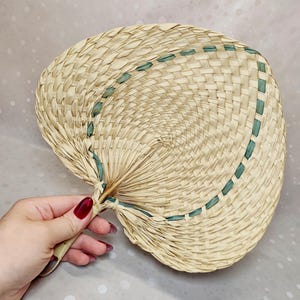 Peut inclure: Un éventail à main fabriqué en paille tressée beige clair, présentant une forme de palette arrondie et une bordure décorative en ruban vert foncé entrelacé sur le bord extérieur de la paille, avec une poignée robuste à la base.