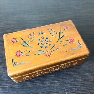 May include: A wooden box with a floral design carved into the top. The flowers are red, blue, and green. The box is made of light brown wood and has a distressed look.