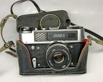 May include: A silver and black 35mm film camera with a leather case. The camera has a lens with the text "Индустар-61 Л/Д" and "2.8/55" printed on it. The camera also has the text "ФЭД 5" printed on it.