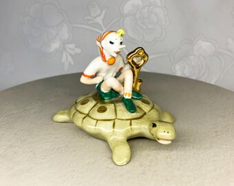 May include: A ceramic figurine of a character with a long nose, wearing a white shirt, green shorts, and a red and orange head covering, sitting on a turtle. The turtle is light green with a brown shell pattern. A gold key is behind the character.