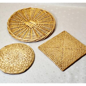 May include: Three woven items including a large round wicker tray with a spiral pattern, a smaller round woven mat with a tight basket weave, and a square woven mat featuring a geometric grid pattern, all in natural light tan colours.