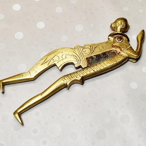 May include: A gold-toned metal nutcracker shaped like a stylized human figure. The tool features intricate engraved scrollwork patterns along the body and limbs, with a dark, textured central section and a small circular accent near the shoulder area.