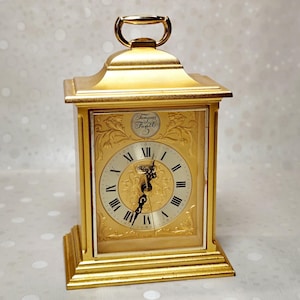 May include: A gold-colored carriage clock with a rectangular frame, a handle on top, and a white circular clock face featuring black Roman numerals. The clock face includes ornate gold filigree detailing and the text Tempus Fugit at the top.