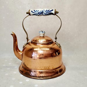 Peut inclure: Une bouilloire à thé en cuivre martelé avec un bec verseur incurvé et un corps arrondi. La bouilloire présente une poignée en céramique blanche et un bouton de couvercle assorti, tous deux décorés d'un motif floral bleu. Fixée par du métal.
