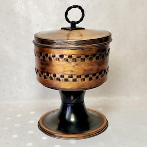 Pode incluir: Um recipiente com tampa na cor cobre, com base de pedestal preta e uma alça circular preta a condizer no topo. O corpo cilíndrico apresenta um padrão axadrezado texturizado e escuro à volta do centro, contrastando com o acabamento metálico.