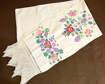 May include: White embroidered table runner with a floral design featuring pink, purple, and green flowers and vines. The runner has a white crocheted lace trim.