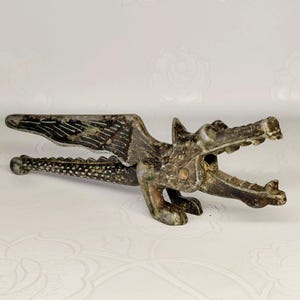 May include: A bronze-colored nutcracker in the shape of a dragon. The dragon has detailed wings and an open mouth. The nutcracker is made of metal and has a textured surface.