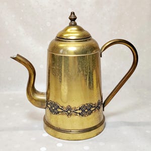 Puede incluir: Una cafetera de latón pulido con un cuerpo cilíndrico alto, un pico curvo y un asa a juego. La base de la cafetera está decorada con una banda oscura de intrincados motivos de volutas, y la tapa está rematada con un pequeño remate puntiagudo.