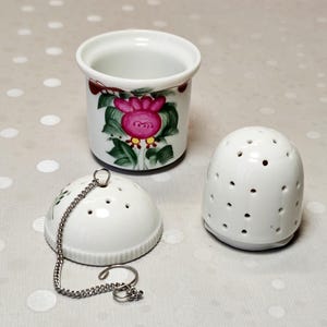 Op de afbeelding: Een witte keramische thee-infuserset bestaande uit een cilindrische beker met een handgeschilderd roze bloem- en groen bladontwerp, een bijpassend geperforeerd koepelvormig deksel en een geperforeerde koepelvormige basis aan een metalen ketting.