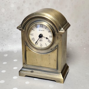 May include: A brass-toned mantel clock with an arched top and a rectangular base. The clock face is off-white with black Roman numerals and the word "INFINITE" printed above the center. It features black hour, minute, and second hands.