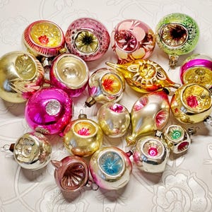 May include: A collection of vintage Christmas ornaments in various shapes and colors. The ornaments include gold, pink, silver, and green glass balls with intricate designs and reflective surfaces. Some have a starburst pattern.