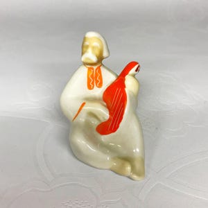 Pode incluir: Uma figurinha de porcelana branca de um homem tocando um instrumento de corda. O homem está vestindo uma camisa branca com detalhes laranja e tem uma barba branca. O instrumento é laranja e vermelho.