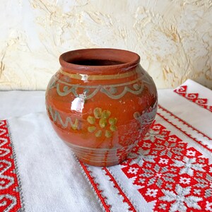 May include: A small, hand-painted terracotta pot with a brown glaze and intricate floral designs in shades of green and yellow. The pot is sitting on a white embroidered tablecloth.