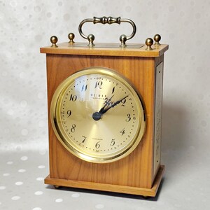 May include: A rectangular wooden mantel clock with a gold-toned circular face featuring black Arabic numerals and the text WEIMAR. The clock has a brass-colored metal handle on top, four small decorative brass finials, and a gold-colored bezel.
