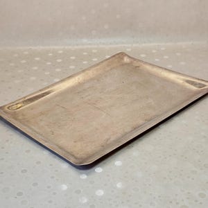 Op de afbeelding: Een rechthoekig dienblad van zilverkleurig metaal met een gladde, platte bodem en licht verhoogde, schuine randen. Het oppervlak van het dienblad heeft een metallic afwerking met subtiele tekenen van slijtage en lichte krassen.