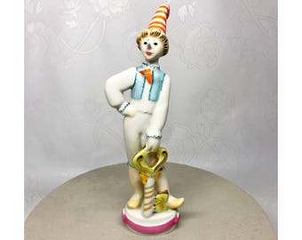May include: A ceramic figurine of a person in a white outfit with a blue vest and an orange and white striped hat. The figurine is holding a large gold key and standing on a pink and white base.