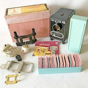 Puede incluir: Un proyector de diapositivas vintage con un cuerpo negro y plateado, una caja de plástico azul y una caja de diapositivas. El proyector está etiquetado como "Экран" y la caja como "Малогабаритный диапроектор "Экран".
