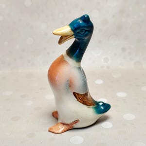 Op de afbeelding: Een keramieken eendbeeldje met een glanzende afwerking, een donkerblauwe kop, een crèmekleurige snavel, een witte borst en bruine en blauwe vleugeldetails. De eend staat rechtop met de snavel licht geopend en heeft een glad, geglazuurd oppervlak over het gehele object.