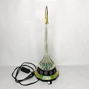 Peut inclure: Une lampe transparente en forme de fusée avec une pointe dorée. La lampe a une base noire avec une bordure verte et jaune. Un cordon d'alimentation noir avec un interrupteur marche/arrêt est connecté à la lampe.