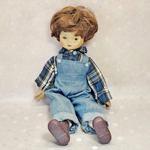 May include: A doll with brown hair and blue eyes wearing a blue plaid long-sleeve shirt under light blue denim overalls. The doll has a pale complexion with freckles and is wearing brown shoes. The doll is seated in a forward-facing position.