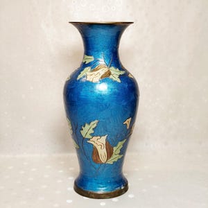 May include: A vibrant blue cloisonné vase featuring a flared rim and a tapered neck. The body of the vase is decorated with light green leaves and cream-colored flower blossoms with brown centers, all outlined in thin, metallic gold-toned wire.