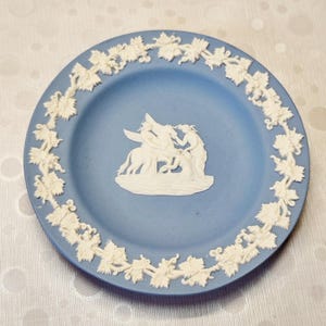 May include: A light blue decorative plate with a white relief design. The plate features a central scene of figures and a border of grapevines. A classic piece for display or serving.
