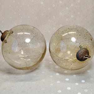 Op de afbeelding: Twee bolvormige glazen ornamenten met een doorschijnende, lichtgouden afwerking en een subtiele, fijn gespikkelde textuur. Elk ornament is voorzien van een decoratieve, geribbelde metalen dop en een klein oogje om het op te hangen.