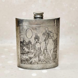 May include: A silver-toned metal flask featuring an embossed scene of two riders on horseback accompanied by a pack of hounds in a field. The flask has a curved rectangular body and a screw-top cap at the centre top, with a blank oval frame.