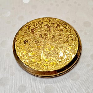 Könnte beinhalten: Ein runder Kompaktspiegel aus goldfarbenem Metall mit einem komplizierten, geprägten Blumen- und Rankenmuster auf der gesamten Oberseite. Das Design besteht aus symmetrischen, wirbelnden Blattmotiven, die von der Mitte des Gehäuses ausgehen.