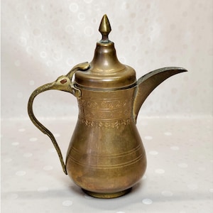 May include: A brass coffee pot featuring a long, curved spout, a handle with a bird-shaped top attachment, and a pointed lid finial. The body is decorated with horizontal bands of etched leaf patterns and simple lines around the midsection.
