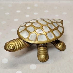May include: A decorative turtle figurine crafted from brass with a polished gold-tone finish. The shell features an intricate inlay of iridescent white mother-of-pearl segments arranged in a geometric pattern, contrasting with the metallic body.