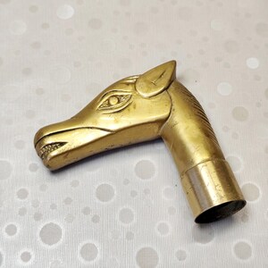May include: A polished brass cane handle shaped like a horse head. The metal piece features detailed engravings for the eye, ear, and mane, with a smooth, curved snout and a cylindrical opening at the base for attachment to a walking stick shaft.