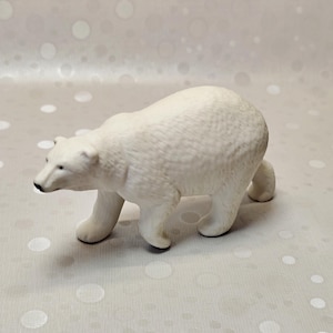 May include: A white polar bear figurine crafted with a textured surface to mimic fur. The bear is depicted in a walking stance with its head lowered, featuring a black nose and small ears. The sculpture is rendered in a matte finish throughout.