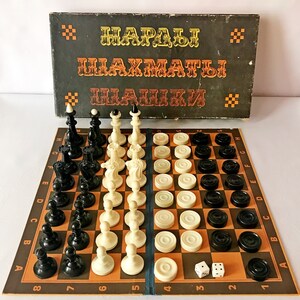 Puede incluir: Un juego de mesa vintage con una caja marrón que dice "Nardy, Ajedrez, Damas" en ruso. El juego incluye un tablero de ajedrez, piezas de ajedrez, damas y un dado.