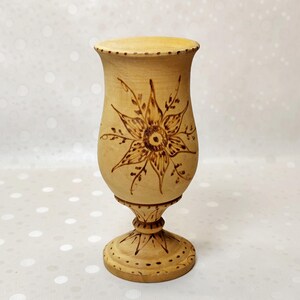 May include: A light tan wooden goblet featuring a dark brown pyrography design of a large, multi-petaled flower on the bowl. The stem and circular base are also decorated with matching dark brown wood-burned patterns and dotted line details.