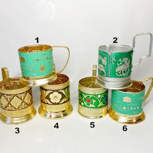 May include: Six different styles of metal tea glass holders. The holders are all gold with green and white designs. The designs include a flower, a geometric pattern, a city skyline, and a floral pattern. The holders are numbered 1 through 6.