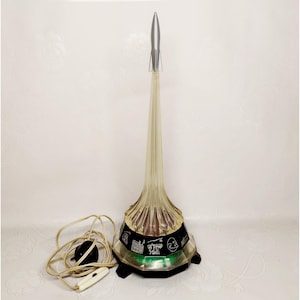 Könnte beinhalten: Eine Vintage-Raketenlampe mit silberner Spitze und einem transparenten, gerippten Körper. Der schwarze Sockel hat ein dekoratives Band mit Illustrationen. Die Lampe hat ein Netzkabel und einen Schalter.