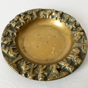 Pode incluir: Um cinzeiro redondo de latão antigo com uma borda em relevo detalhada. A borda apresenta uma série de figuras e animais esculpidos. O interior do cinzeiro tem uma superfície dourada lisa. É um item vintage.