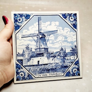 May include: A square ceramic tile featuring a blue and white illustration of a windmill. The scene depicts the De Valk windmill in Montfoort with clouds in the background and floral corner patterns. The text De Valk, Montfoort is printed.
